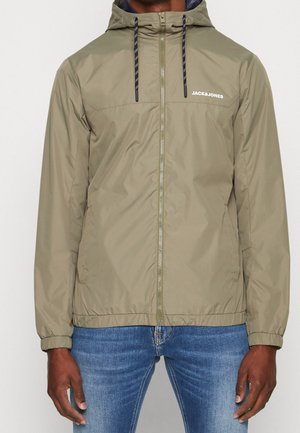 Man draagt een licht olijfgroene capuchontrui met rits, zwarte trekkoorden en "JACK&JONES" logo, gecombineerd met blauwe spijkerbroek.