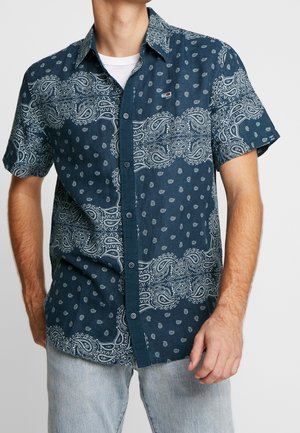 Homme portant une chemise à manches courtes bleu marine à boutons avec des motifs cachemire blancs et de petits points, associée à un jean bleu clair.