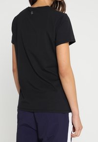 Mujer vestida con una camiseta negra de manga corta lisa y pantalones cortos azul oscuro con franjas laterales blancas, vista desde la espalda contra un fondo blanco.