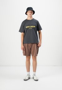 Tânăr purtând o pălărie tip bucket neagră, un tricou grafic gri închis, șorturi cargo maro, șosete albe și adidași albi, pe un fundal simplu.