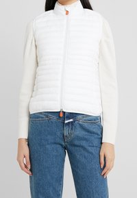 White padded vest with a high collar, front zipper, and orange zipper pull; worn over a cream sweater, paired with blue jeans.