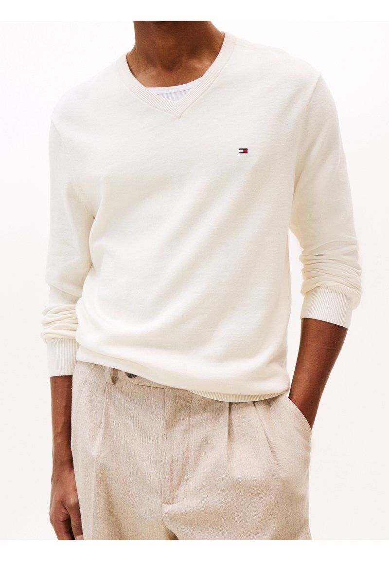Homme portant un pull col en V écru avec un petit logo drapeau et un pantalon beige plissé, mains dans les poches sur fond uni.