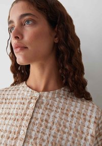 Chemise légère à motifs beige et blanche avec un col rond et une fermeture par bouton, présentant un design en tissu texturé à carreaux.