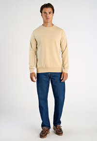 Beige puuvillapaita, jossa on pyöreä pääntie, röyhelöhiha ja helma. Yhdistetty sinisiin farkkuihin ja ruskeisiin venetsialaisiin kenkiin. Valkoinen teksti rinnassa.