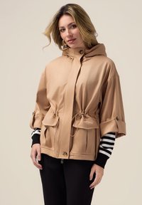 Lysebrune hættejakke med snøre i taljen, store frontlommer og optrukne ærmer. Glat tekstur og subtil glans.