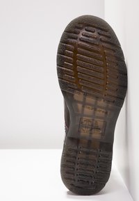 Semelle d'une botte brune avec motif de crampons rainurés, marquage en relief et un logo avec texte sur la zone du talon, photographiée sur un fond blanc.