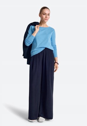 Žena v svetlomodrom tričku s dlhým rukávom a širokých tmavomodrých nohaviciach drží tmavomodrý kabát na pleci, má na nohách biele topánky a tmavý náramok.