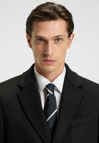 Cappotto di lana nero con colletto classico, abbinato a una camicia bianca e una cravatta nera con sottili righe bianche. Tessuto liscio e strutturato.
