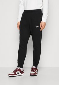 Osoba pohybující se v černých Nike teplácích a bílých, bordo a černých Nike teniskách, stojící na jednobarevném bílém pozadí.
