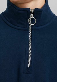 Sudadera con cremallera en navy y cuello alto, que presenta un cierre de cremallera plateado y un tirador en forma de anillo, fabricada con una tela suave y texturizada.