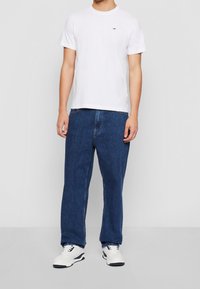 Vit rundhalsad T-shirt med liten logotyp, matchad med blå baggy jeans och vita sneakers med svarta detaljer. Enfärgad bakgrund.