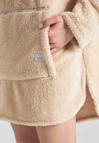 Beżowy szlafrok z polaru o miękkiej teksturze. Posiada przednią kieszeń i naszywkę. Projekt obejmuje otwarty przód i krótki krój.