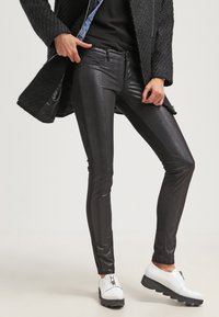 Zwarte synthetische leren broeken met een glanzende afwerking, slim fit en voorzakken, gecombineerd met witte rits schoenen en een gestructureerde zwarte jas.