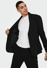 Blazer nero con un solo bottone, due tasche e rivestimento in raso. Indossato sopra una maglietta bianca, mette in risalto una vestibilità elegante e un design moderno.