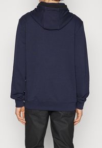 Felpa blu navy con tasca a marsupio, cappuccio con cordino e polsini a coste; mostrata con una persona di spalle, che indossa pantaloni neri.