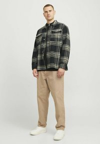 Jachetă de flanel în carouri alb-negru cu două buzunare pe piept, purtată peste un top negru. Pantaloni bej și adidași albi completează ținuta.