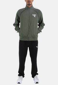 Tuta sportiva con giacca verde con cerniera e dettagli bianchi, abbinata a pantaloni neri. Realizzata in tessuto morbido, con dettaglio logo.