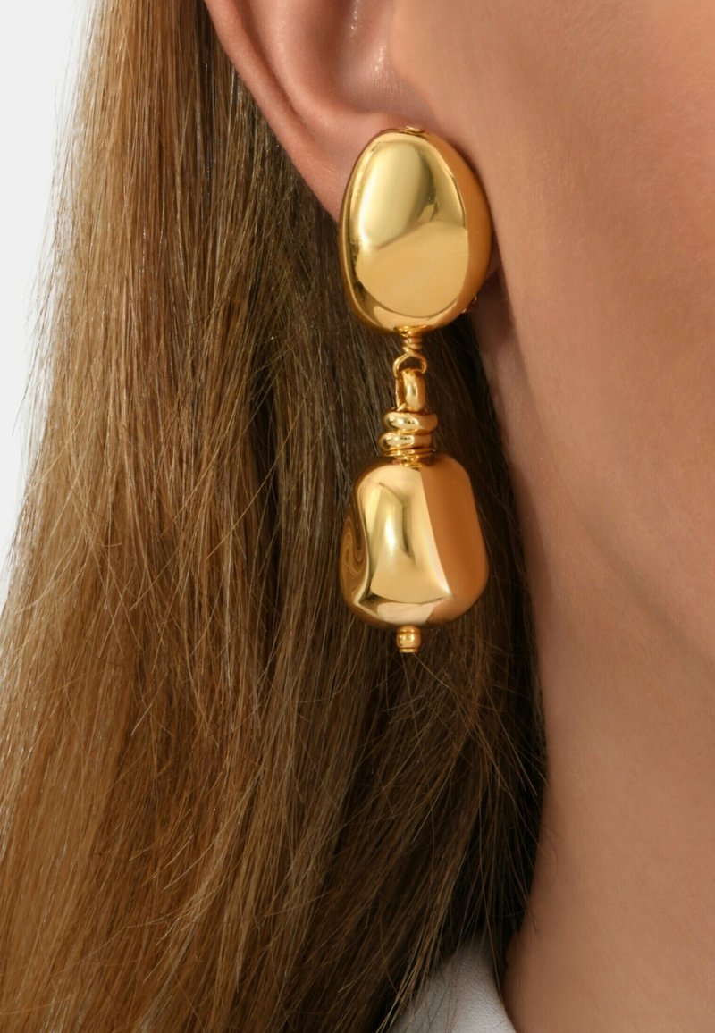 Gros plan sur une boucle d'oreille en forme de goutte irrégulière de couleur dorée, portée sur l'oreille d'une femme aux cheveux châtain clair.