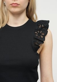 Γυναίκα που φοράει μαύρο τοπ χωρίς μανίκια με ριμπ ύφασμα και διακοσμητικά από βολάν δαντέλας με λουλουδένιο σχέδιο στον δεξιό ώμο.