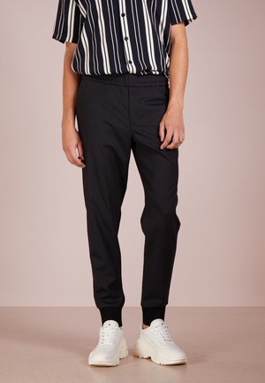 Mężczyzna ubrany w czarne spodnie dresowe, białe masywne trampki oraz czarno-białą koszulkę w pionowe paski z krótkim rękawem na beżowym tle.