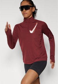 Bordo dugi rukav sportski majica s visokim ovratnikom i zipom. Sadrži bijeli swoosh logo i otvore za prste na rukavima, u kombinaciji s crnim šortsevima.