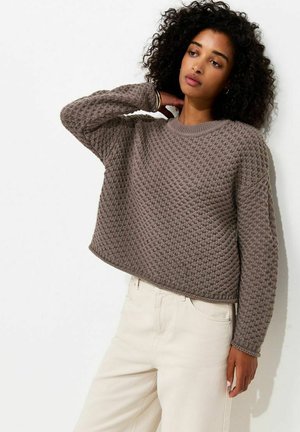 Donna con capelli ricci che indossa un maglione marrone a maglia con trama e pantaloni beige chiaro, posando con una mano dietro al collo.
