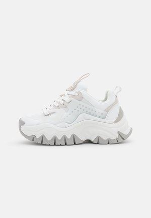 Hvid chunky sneaker med tyk bølget grå sål, snøredesign, mesh tunge og trækflige på tunge og hæl mod ensfarvet baggrund.
