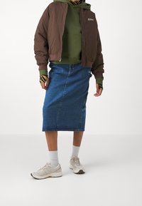 Osoba oblečená v hnědém bombardéru přes zelenou mikinu, modré džíny sukně, bílé ponožky a béžové tenisky na čistém bílém pozadí.