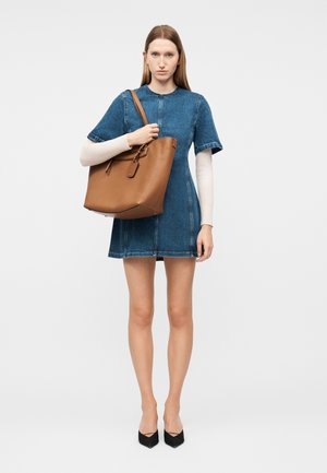 Γυναίκα με κοντομάνικο φόρεμα από τζιν που κρατά μια μεγάλη καφέ δερμάτινη τσάντα, φοράει μαύρες γόβες και λευκό μακρυμάνικο τοπ από κάτω.