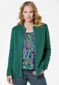 Groene textuur cardigan met een ronde hals, rits aan de voorkant en een enkele zak, gelaagd over een kleurrijke gestreepte top en donkere spijkerbroek.