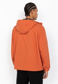 Oranje hoodie-jack met een soepele textuur, met een ontspannen pasvorm, elastische manchetten en subtiele stiksels bij de schouders.