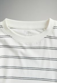 Camiseta blanca de algodón con rayas horizontales negras. Presenta un escote redondo y una etiqueta que dice "THE SET", talla M, hecha en Bangladesh.