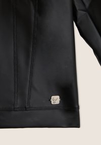 Garment in tessuto elasticizzato nero con cuciture a vista e accentuazione del logo metallico. Texture liscia e design aderente.