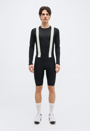 Joven de pie, vestido con una camiseta térmica de manga larga negra para ciclismo, culotte con tirantes blancos, calcetines blancos y zapatillas de ciclismo blancas.