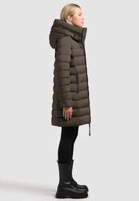 Płaszcz puchowy w ciemnym brązie z poziomym pikowaniem, wysokim kołnierzem i kieszeniami na zamek. Połączony z czarnymi legginsami i sznurowanymi botkami.