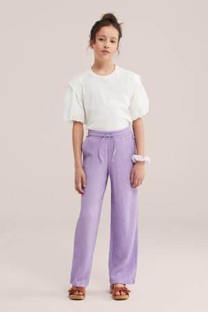 Fiatal személy, aki kontyba fogta a haját, fehér puffos ujjú felsőt, texturált levendula színű, zsinóros nadrágot, szandált visel, és fehér hajgumikat a csuklóján.