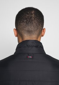 Schwarze isolierte Jacke mit hohem Kragen, die horizontale Nähte und ein kleines Logo-Emblem in Rot, Weiß und Blau auf dem Rücken hat.