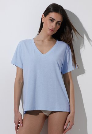 Donna con capelli lunghi e scuri che indossa una maglietta azzurro chiaro a maniche corte con scollo a V, in piedi contro un muro bianco con un'ombra proiettata dietro.