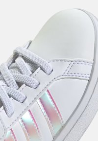 Vita lädersneaker med tre iriserande ränder, grå snören och en texturerad gummisula. Detaljstickning längs kanterna.