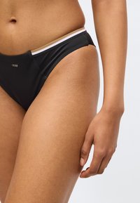 Melnas bikinu apakšdaļas ar kontrastējošu jostas joslu, kurā ir baltas un zelta svītras. Gluda tekstūra, piegulošs dizains un minimāla zīmola izcelšana.