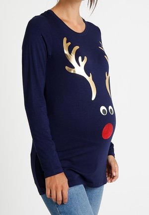 Zwangere vrouw die een marineblauw shirt met lange mouwen draagt met gouden rendiergeweien, witte ogen en een rood neusontwerp.