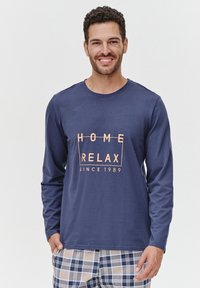 Granatowa koszula z długim rękawem z napisem "HOME RELAX SINCE 1989" w kolorze brzoskwiniowym, zestawiona z piżamowymi spodniami w kratę w odcieniach niebieskiego, pomarańczowego i białego.