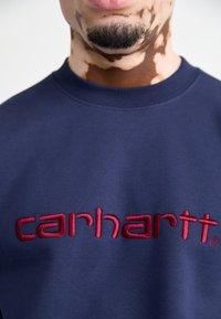 Sudadera Navy hecha de una tela suave, con el logotipo "carhartt" bordado en rojo. Cuello redondo y puños de punto.