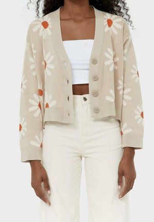 Femme portant un cardigan beige boutonné avec des motifs de marguerites blanches et orange, un haut tube blanc et un pantalon écru taille haute.