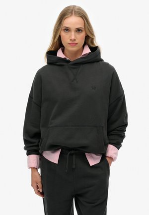 Sudadera gris oscura de gran tamaño con un bolsillo canguro, capucha con cordón y un logotipo sutil. Llevada sobre una camisa color rosa claro con cuello.