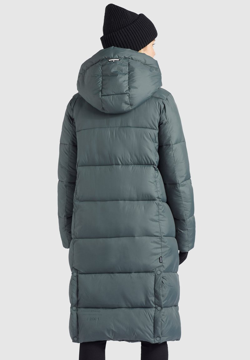 Person, der einen langen, dunkelblauen gepolsterten Mantel mit Kapuze und eine schwarze Strickmütze trägt, steht mit dem Rücken zu einem schlichten hellen Hintergrund.