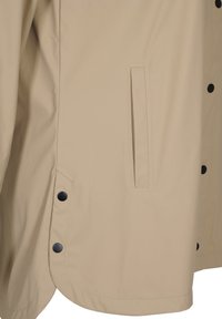 Section de veste beige montrant une poche fendue verticale et des boutons-pression noirs sur le côté et l’ourlet avant.