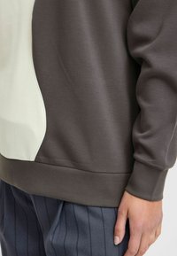 Brown relaxed-fit sweatshirt with a contrasting cream side panel; soft texture, ribbed cuffs, and a straight hem. Paired with pinstriped trousers.