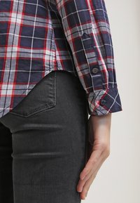 Červeno-bílý a modrý kostkovaný košile s dlouhými rukávy a knoflíkovými manžetami, v kombinaci s tmavě šedými přiléhavými džíny, které ukazují textury a vzory tkanin.