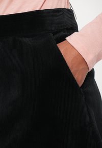 Svarte corduroy-bukser med et teksturert ribbemønster og sidelomme, kombinert med en lys rosa langermet skjorte.